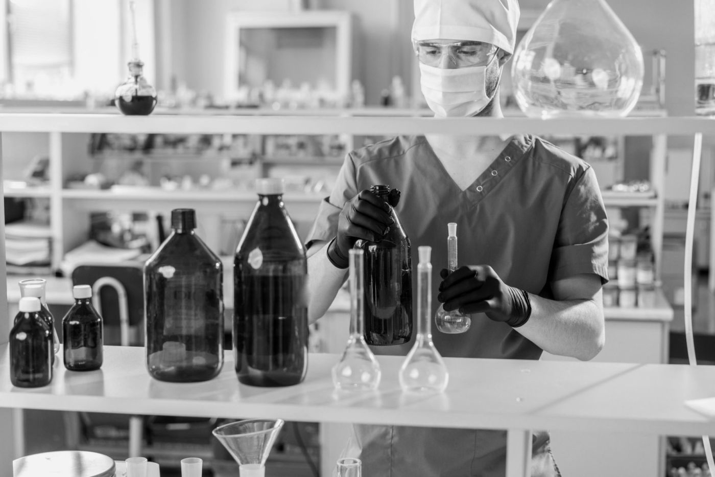 Technician in a chemical laboratory handling dark glass bottles and flasks, black and white photograph